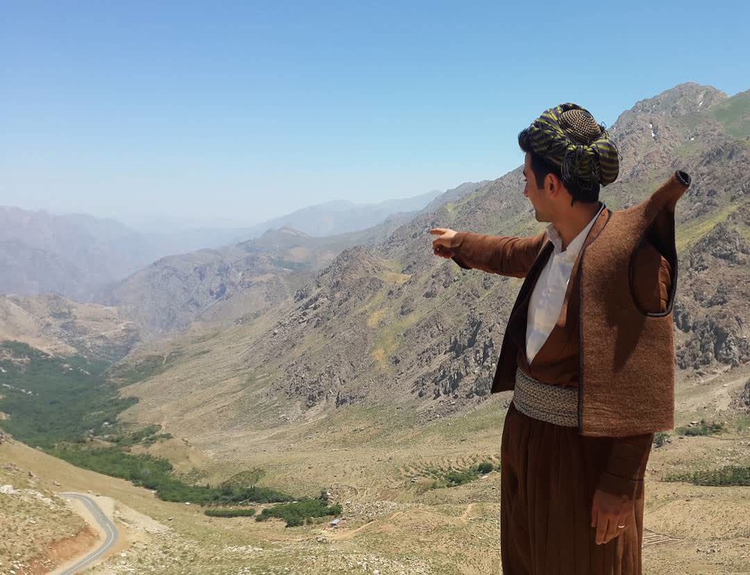 Kahverengi keçe yelek (faranji), şûtik kuşak ve çizgili puşi sarığıyla dağ manzarasını gösteren Hawrami erkeği
