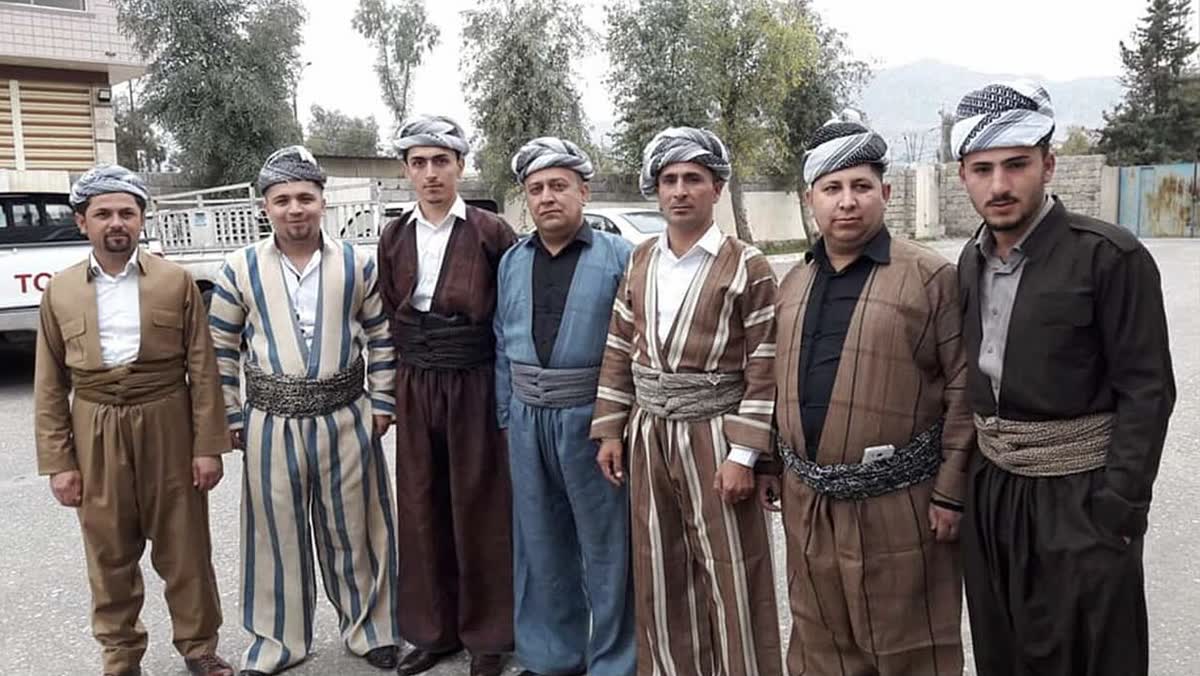 Kahve, mavi, bej ve çizgili tonlarda şal û şepik takımları giymiş yedi adam yan yana