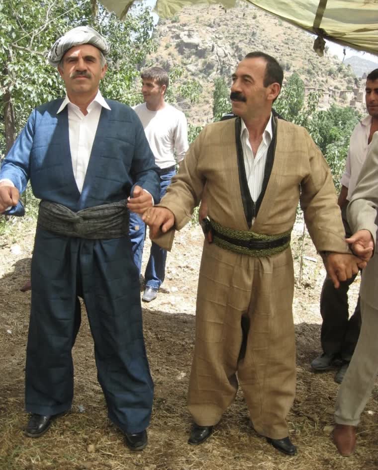 Açık havada düğünde şal û şepik takımları giymiş adamlar el ele halay çekiyor