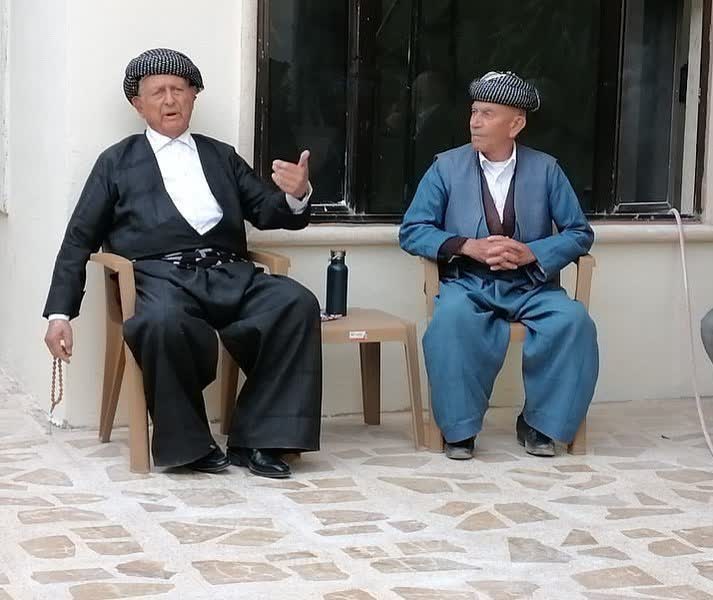 Gri çizgili şal û şepik takımları giymiş, puşi sarıklı iki yaşlı beyefendi sohbet ederken