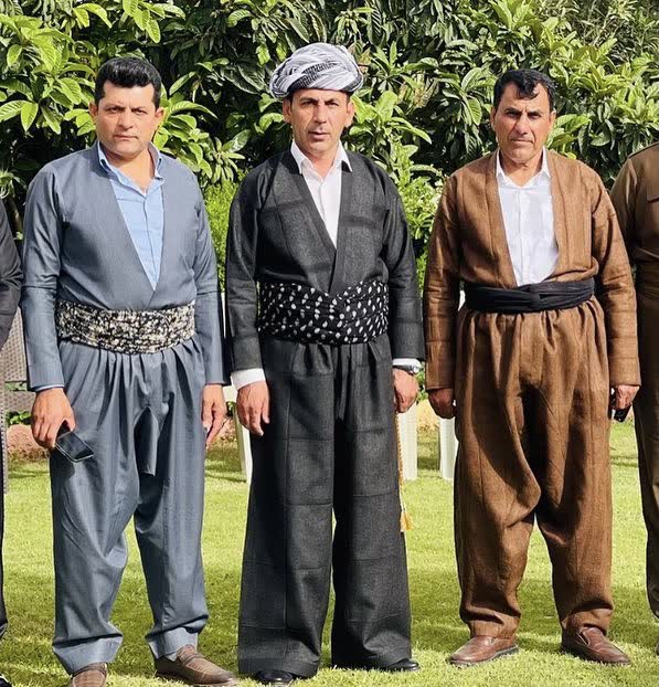 Lacivert, kahve ve gri şal û şepik takımları giymiş üç adam bahçede yan yana